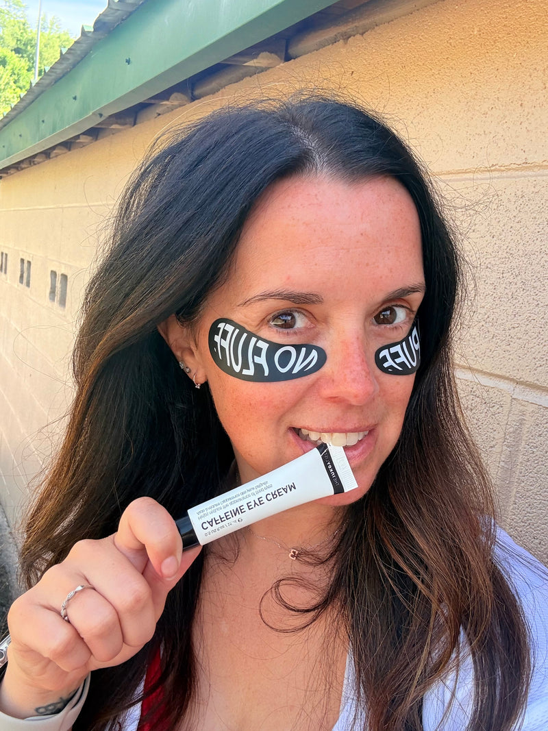 A woman with black and white under eye patches holding up a caffeine eye cream tube