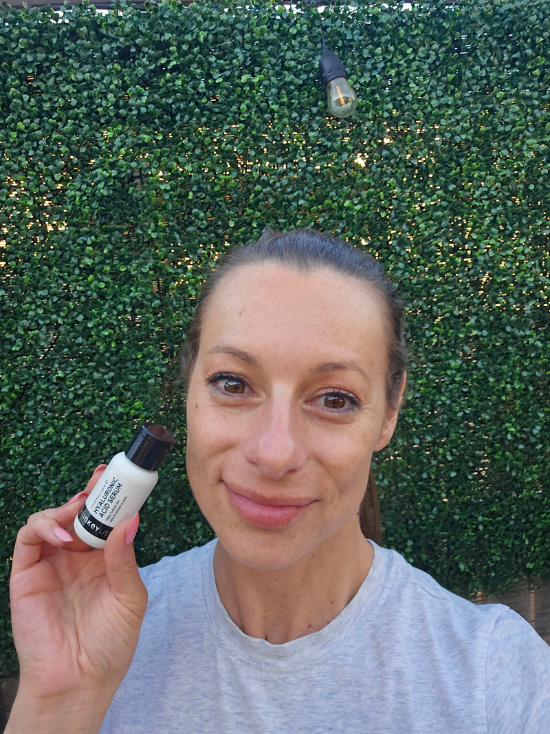 A woman holding a bottle of Hyaluronic Acid Serum to her face against a green background