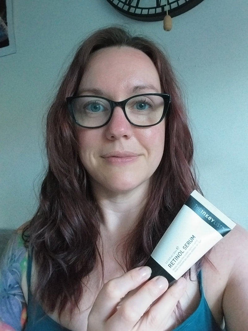 A woman with long reddish-brown hair and black-rimmed glasses holds a white tube of Retinol Serum in front of her face, set against a domestic background with a wall clock.