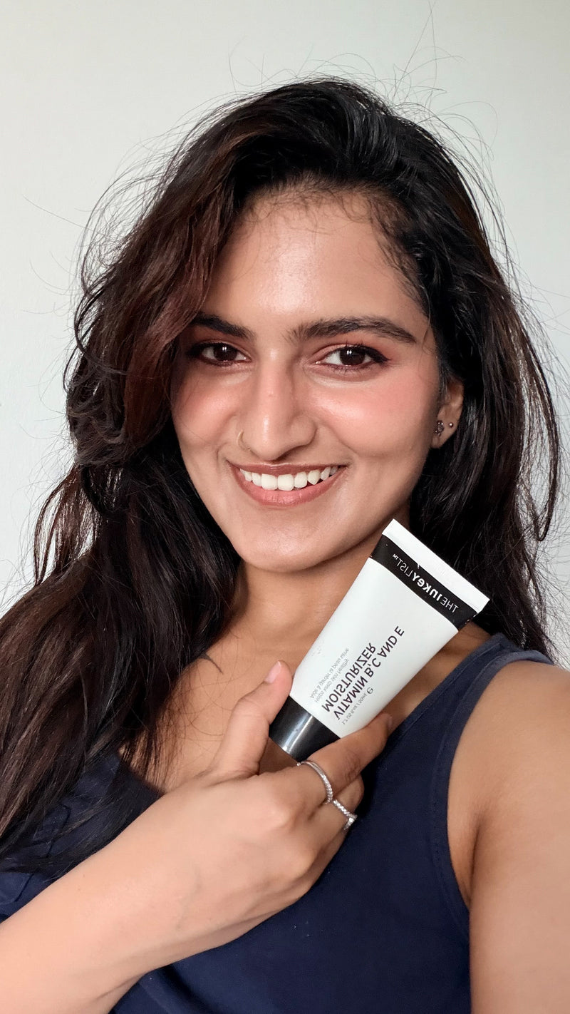 A smiling woman with long dark hair holds up a white and black tube of Moisturizer against a plain light gray background.