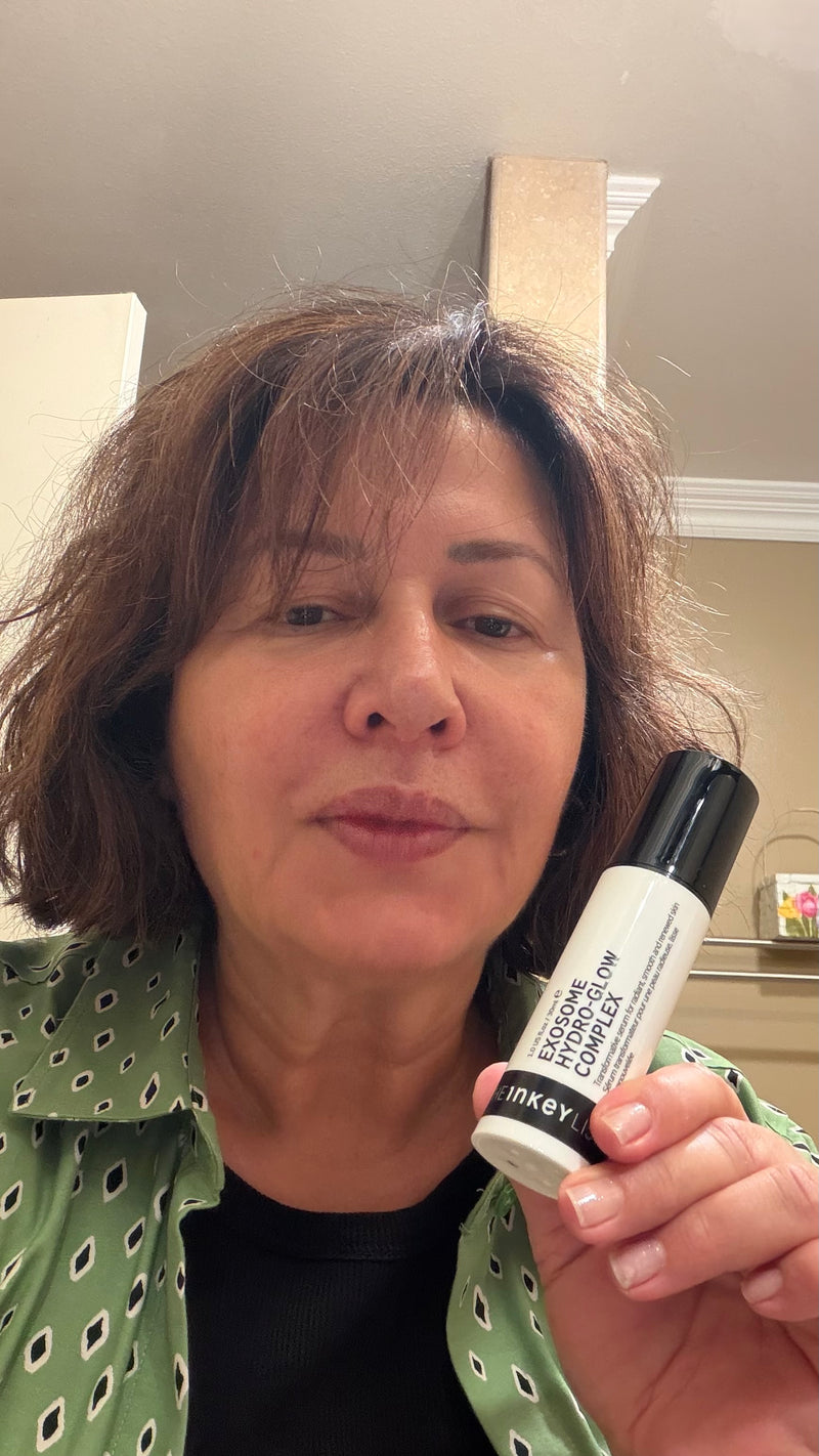 A woman holding a white and black Exosome Serum in front of her face, set against an indoor home background.