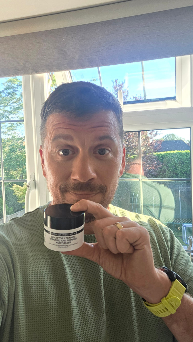 A man holding a small white cosmetic jar with a black lid up to his face, taken in a selfie-style indoor photo with a window and outdoor greenery in the background.