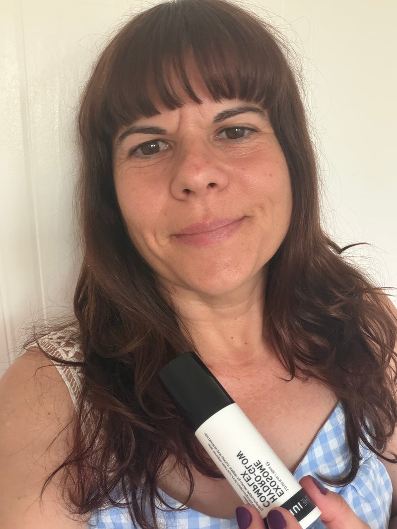 A woman with brown hair holding a white and black and white bottle of Exosome Glow Serum against a plain off-white wall background.
