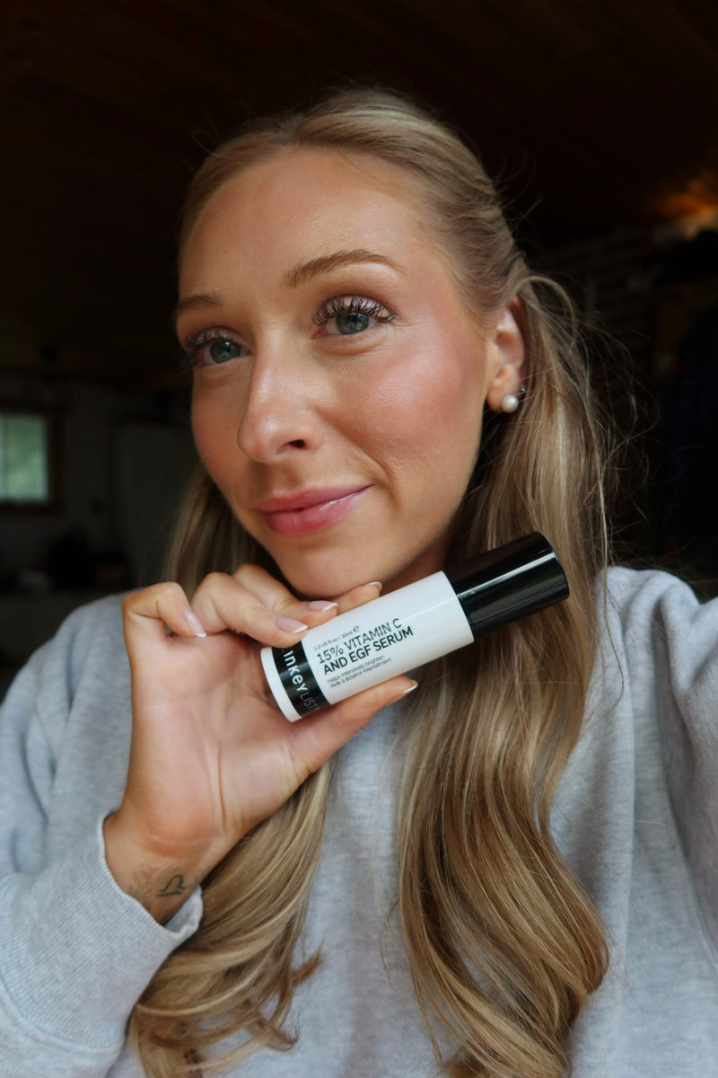 A woman with blonde hair holding a white and black tube of Serum, wearing a light gray sweatshirt in an indoor setting.