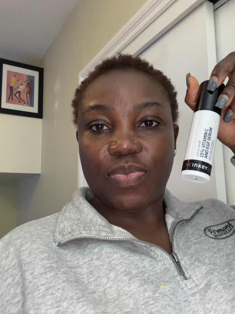 A woman holding a white and black Inkey 15% Vitamin C Serum bottle in front of her face, wearing a gray zip-up sweatshirt in an indoor setting.