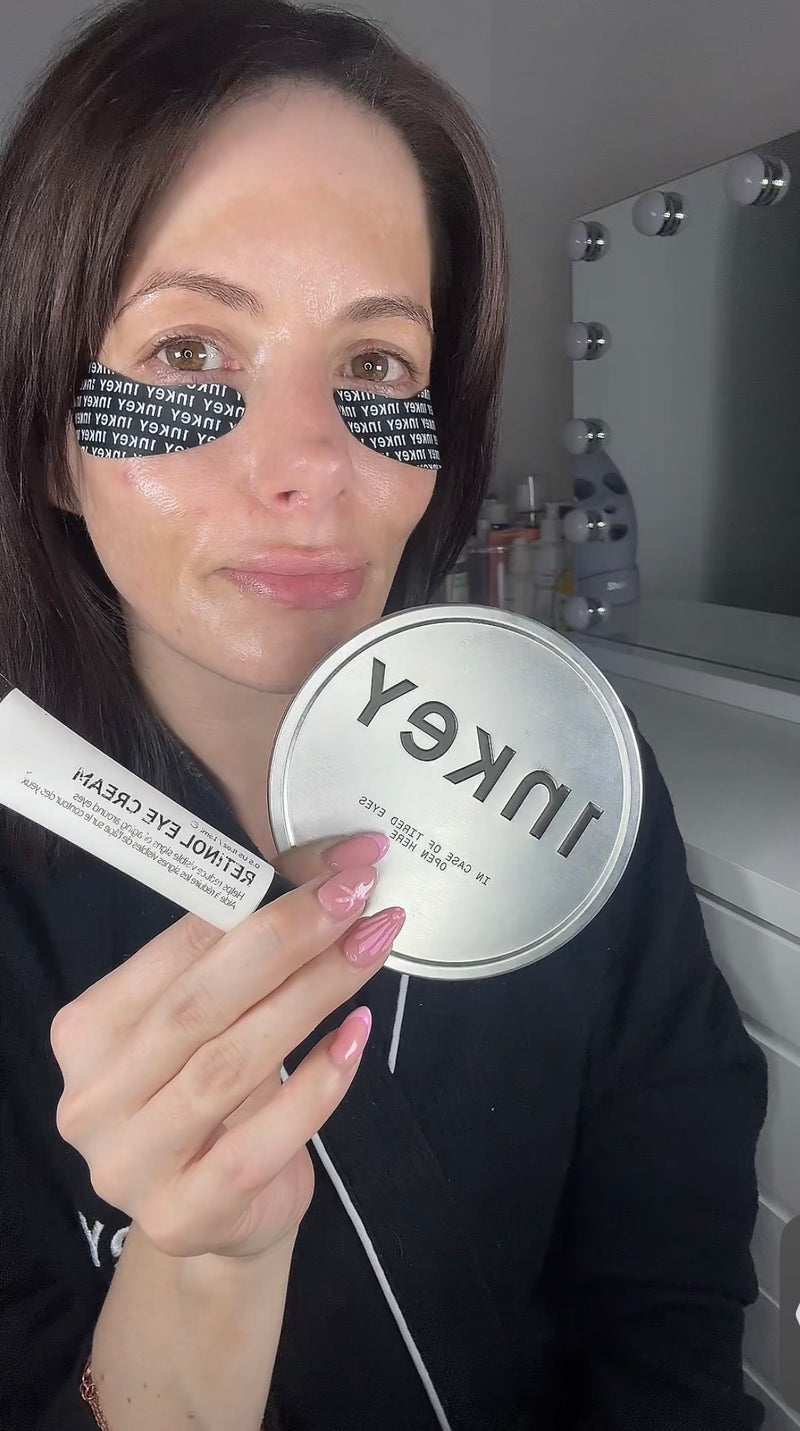 A woman holding a round tin and a white tube of Inkey Retinol Eye Cream in front of her face, with black eye patches on her eyes and a vanity mirror in the background.