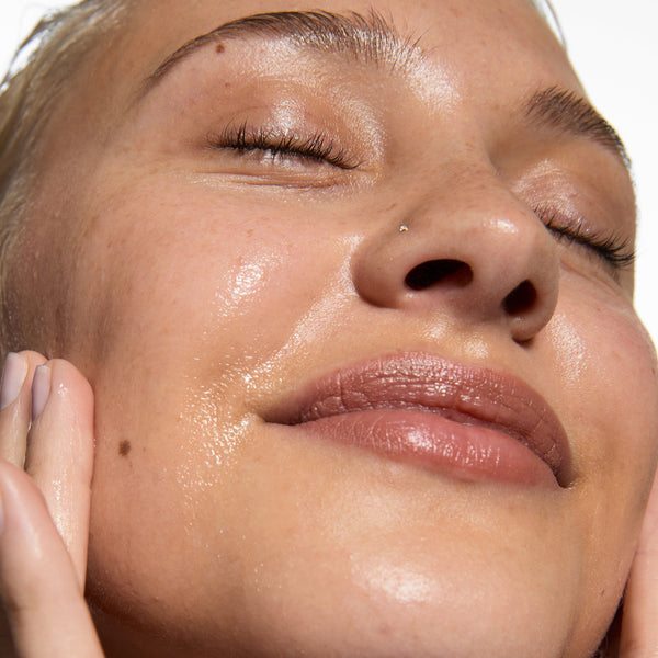 Close-up of a woman applying or blending a cream makeup product on her cheek, with her hand near her face against a plain white background.