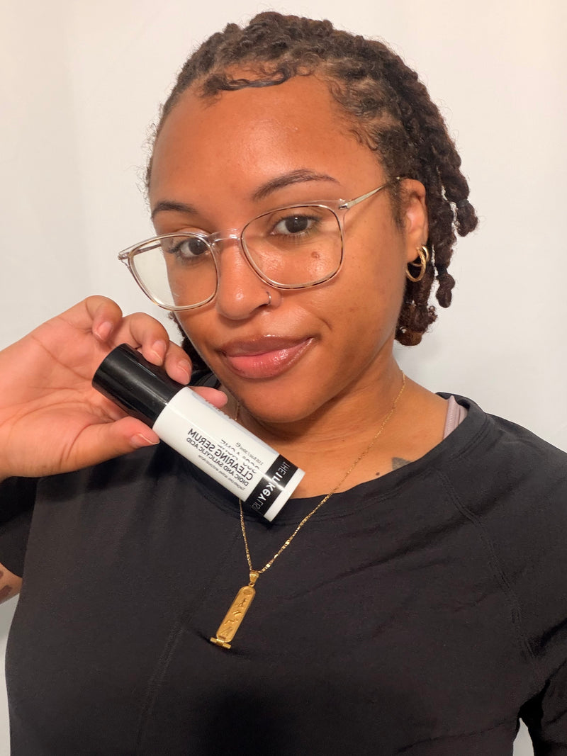 A woman with glasses and braided hair holds a black and white bottle of '360 Serum' next to her face, wearing a black shirt and a gold pendant necklace against a plain light background.