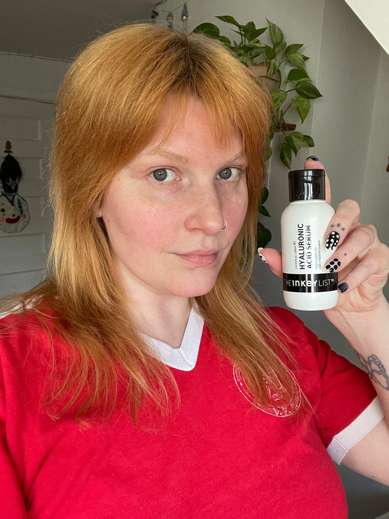 A woman with red hair in a bathroom setting holding up a bottle of hyaluronic acid serum