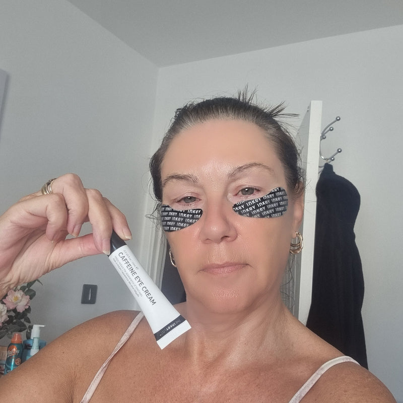 A woman holding a white tube of caffeine eye cream in front of her face, with black eye patches.