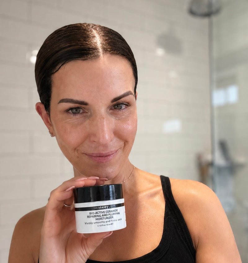 A woman in a black tank top holds a jar of Bio active moisturiser in a bright bathroom setting with white tiles.