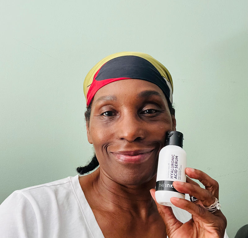 A woman wearing a black, yellow, and red headband holds up a white bottle of The Inkey List Hyaluronic Acid Serum' against a light mint green wall.