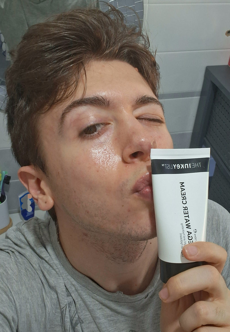 A man in a gray t-shirt holding a white and black tube of Omega Water Cream to his lips in a bathroom setting.
