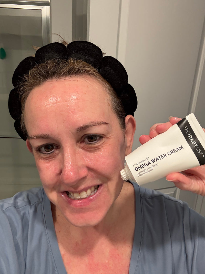 A woman wearing a black scrunchie headband and blue scrub top holds up a white tube of 'The Inkey List' Omega Water Cream in a bathroom setting.