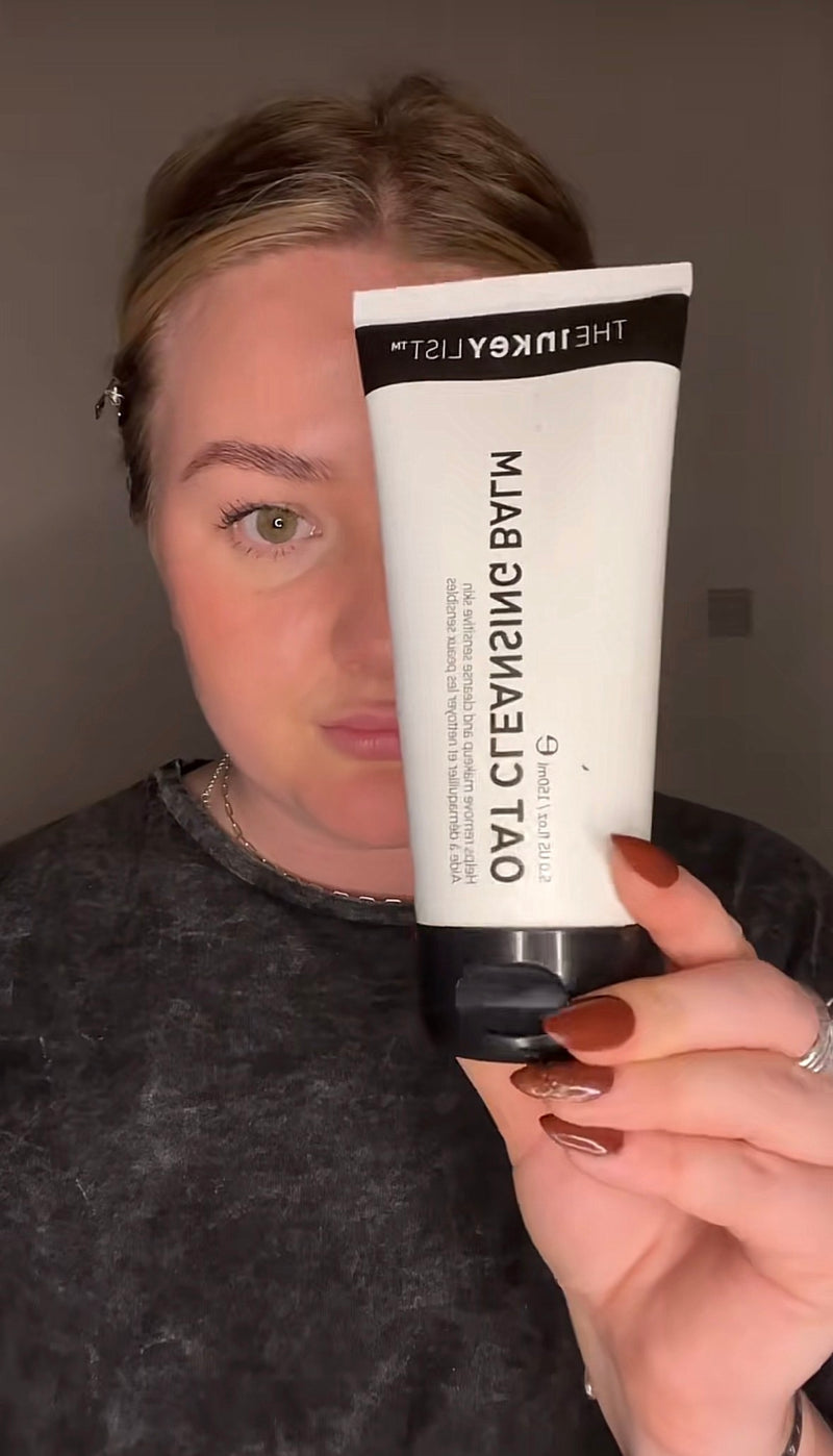A woman holds up a white tube of The Inkey List Oat Cleansing Balm in front of her face, showing the product in a close-up shot against a plain gray wall.