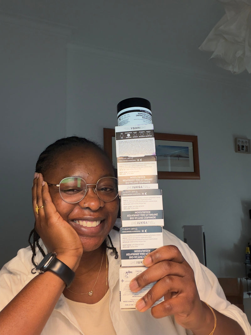 A woman with glasses and braids holds up a tall stack of white product boxes in a home setting with a gray wall and framed picture in the background.