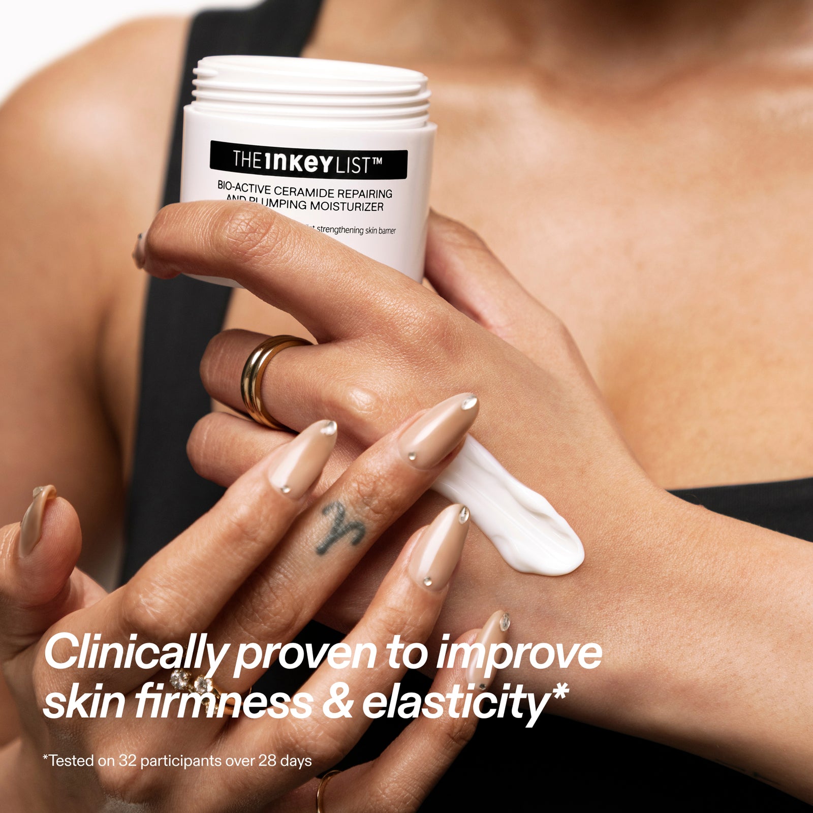 A close-up of a person applying white cream from The Inkey List Bioactive Repairing Moisturizer onto their hand. The person is wearing beige manicured nails and gold rings, and the background is a soft-focus view of their bare torso.