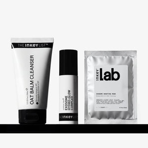 Three skincare products from The Inkey List arranged on a black shelf against a light gray background: a oat Balm Cleanser tube, an Exosome Complex Flow cream bottle, and an Exosome Boosting Mask sachet.