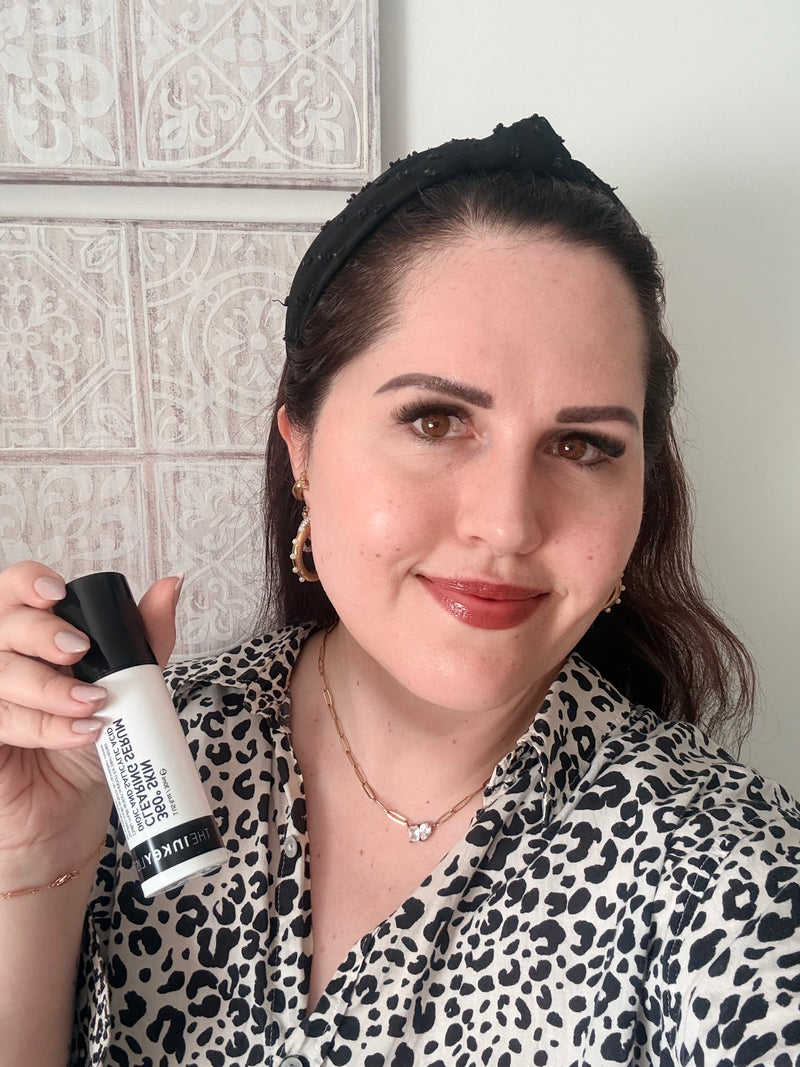 A woman wearing a black headband and a black-and-white leopard print top holds up a bottle of Inkey 360 Serum in a mirror selfie style. The background features a white wall and decorative gray tiles.