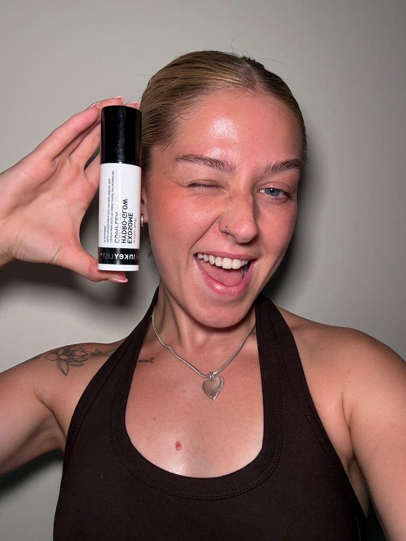 A woman holding up a white and black bottle of Exosome Glow Serum against a plain gray wall.