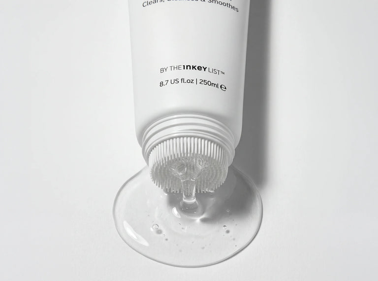 A white squeeze tube of hair product lies on its side with clear gel or serum dripping from the nozzle onto a white surface. The tube shows the text 'BY THE INKEY LIST' and 'Clean, Smoothed & Smoothies'.