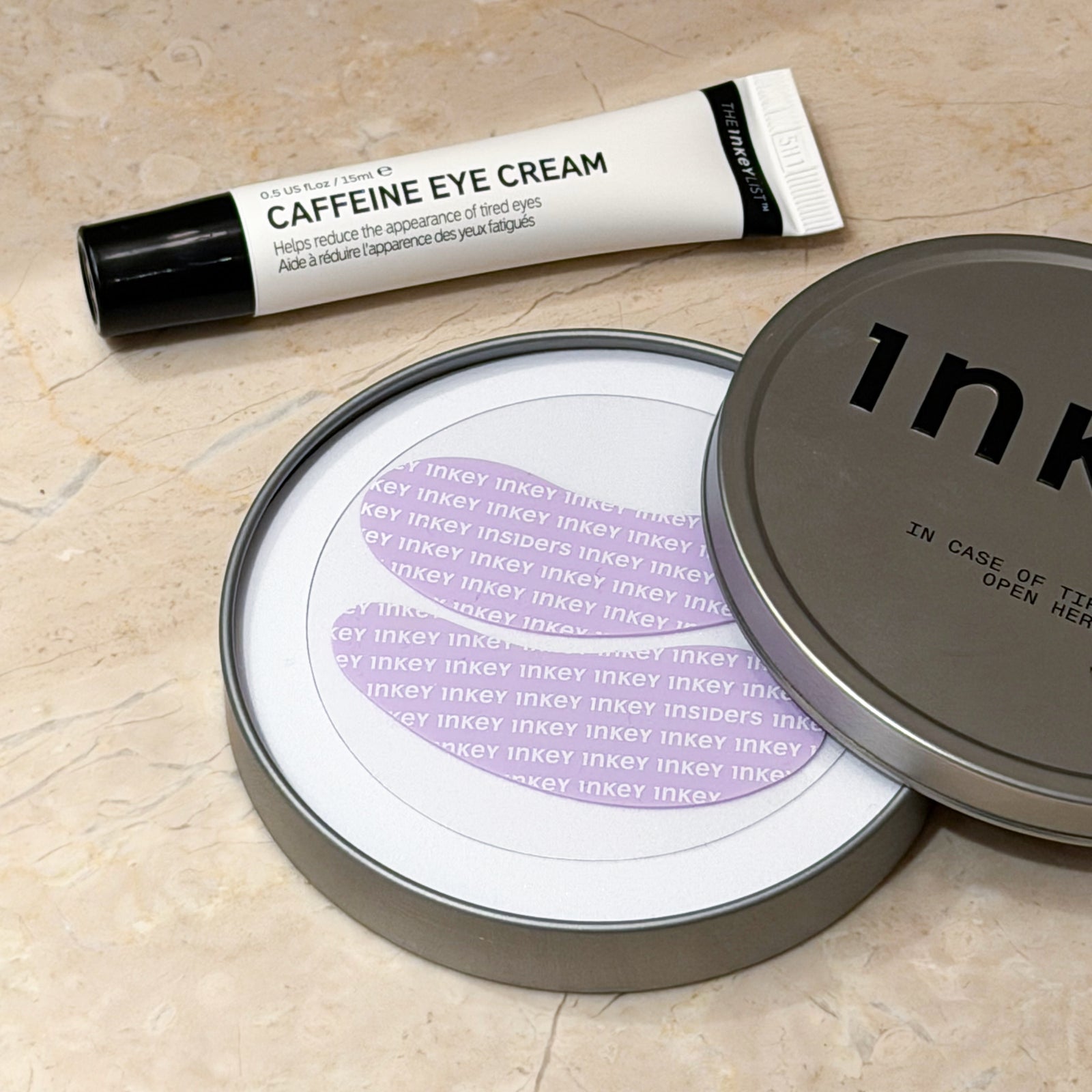 A skincare set featuring a tube of caffeine eye cream and an open round metal tin of Inky Ink eye insitters on a beige marble surface.