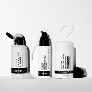 Three white skincare containers from The Inkey List with black caps and black text, arranged on a white surface against a white background. A black applicator lies on the left, a pump bottle stands in the center, and a stick container with a white cream smear above it stands on the right.