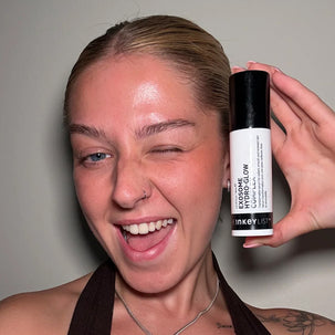 A woman winking while holding up a bottle of InkeyList Exosome HydroGlow Complex against a plain gray wall.