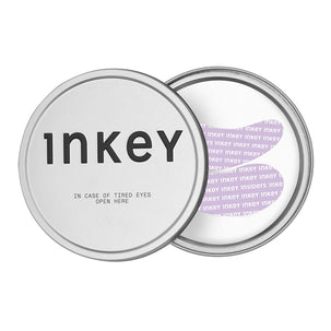 Two round silver metal tins for Inkey cosmetics, one closed with black text reading 'inkey' and 'IN CASE OF TIRED EYES OPEN HERE', and one open showing a light purple patterned interior with the brand name repeated.