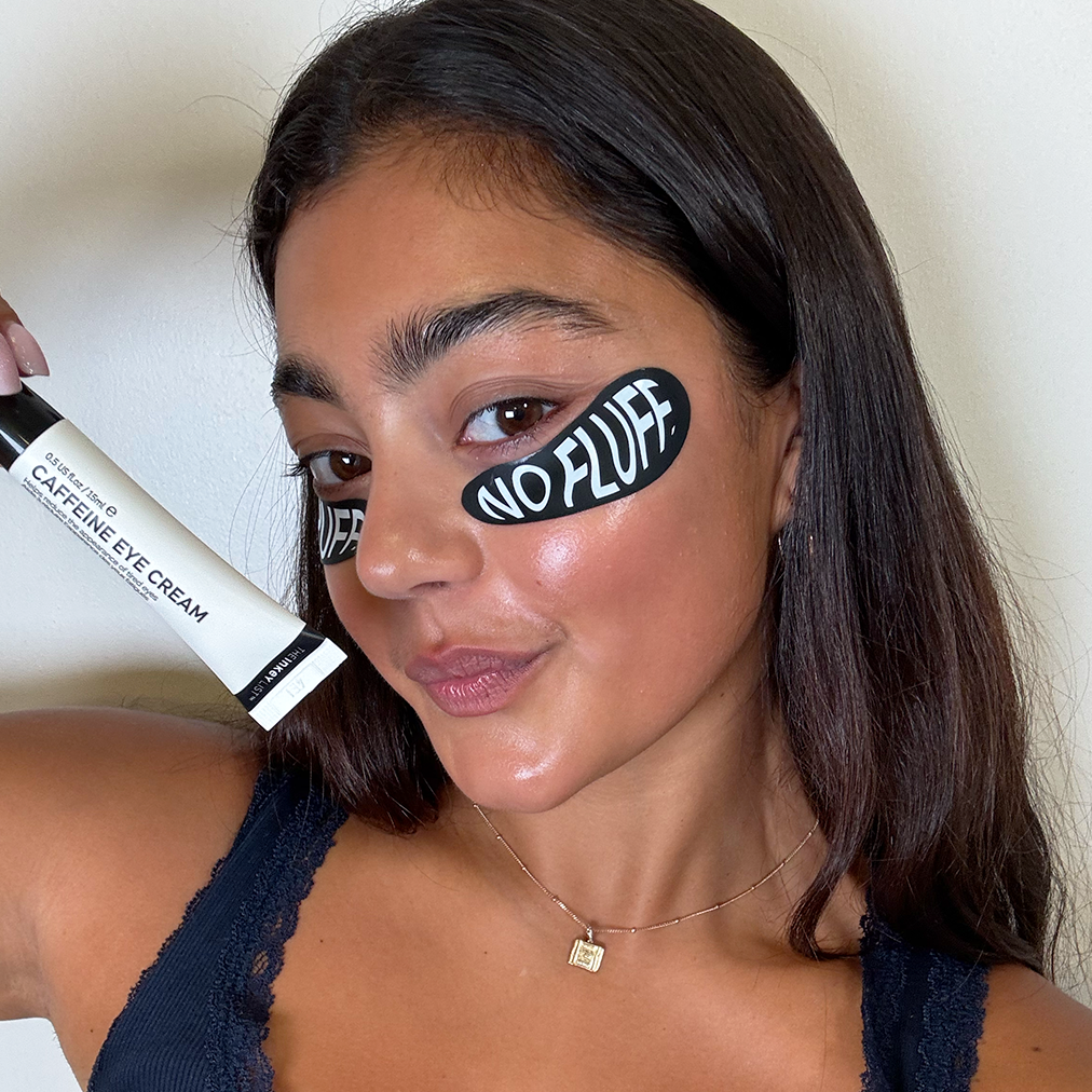 Woman holding a tube of caffeine eye cream with 'NO FLUFF' eye patch under her eye