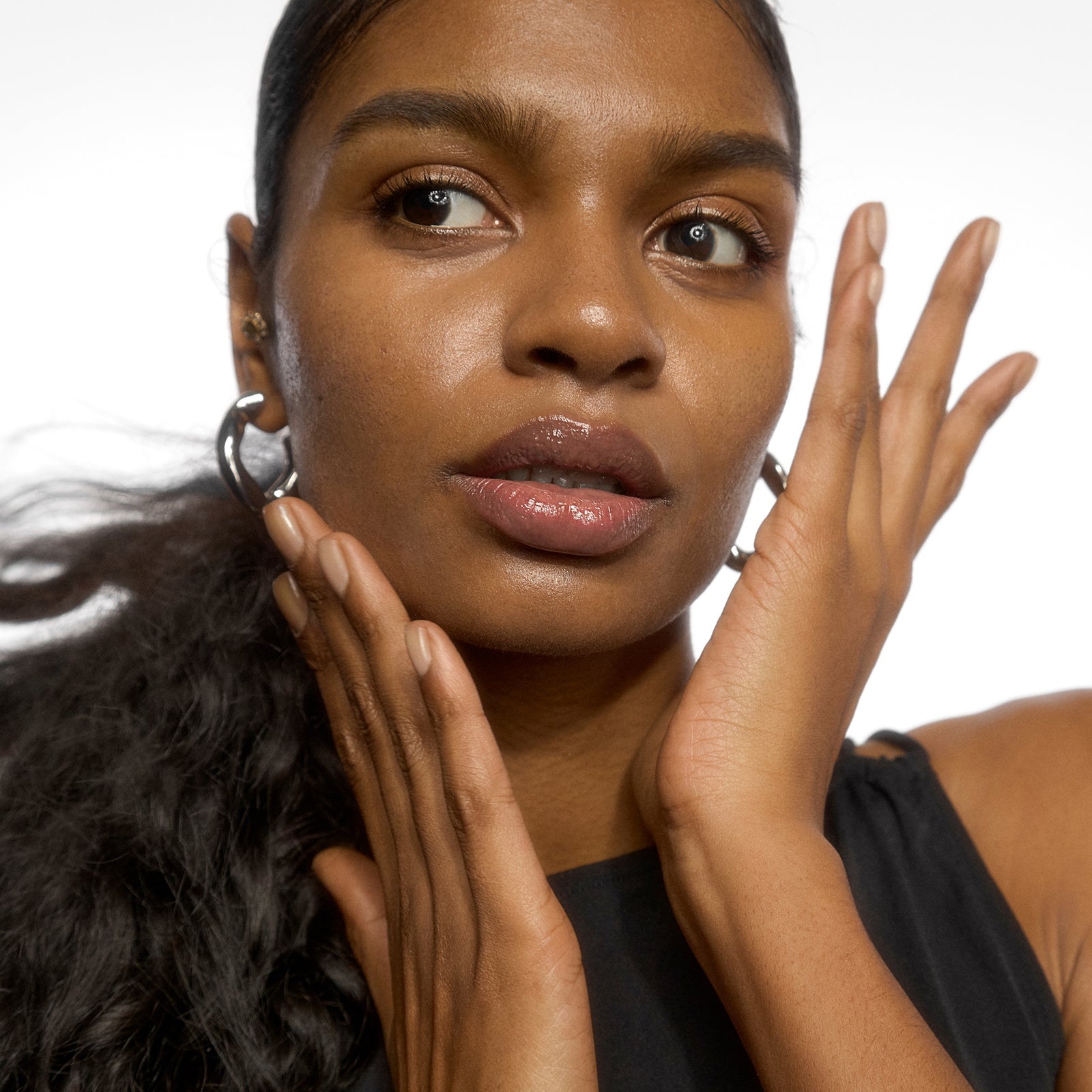 A close-up portrait of a woman with dark curly hair wearing silver hoop earrings and a black sleeveless top, posing with her hands near her face against a plain white background.