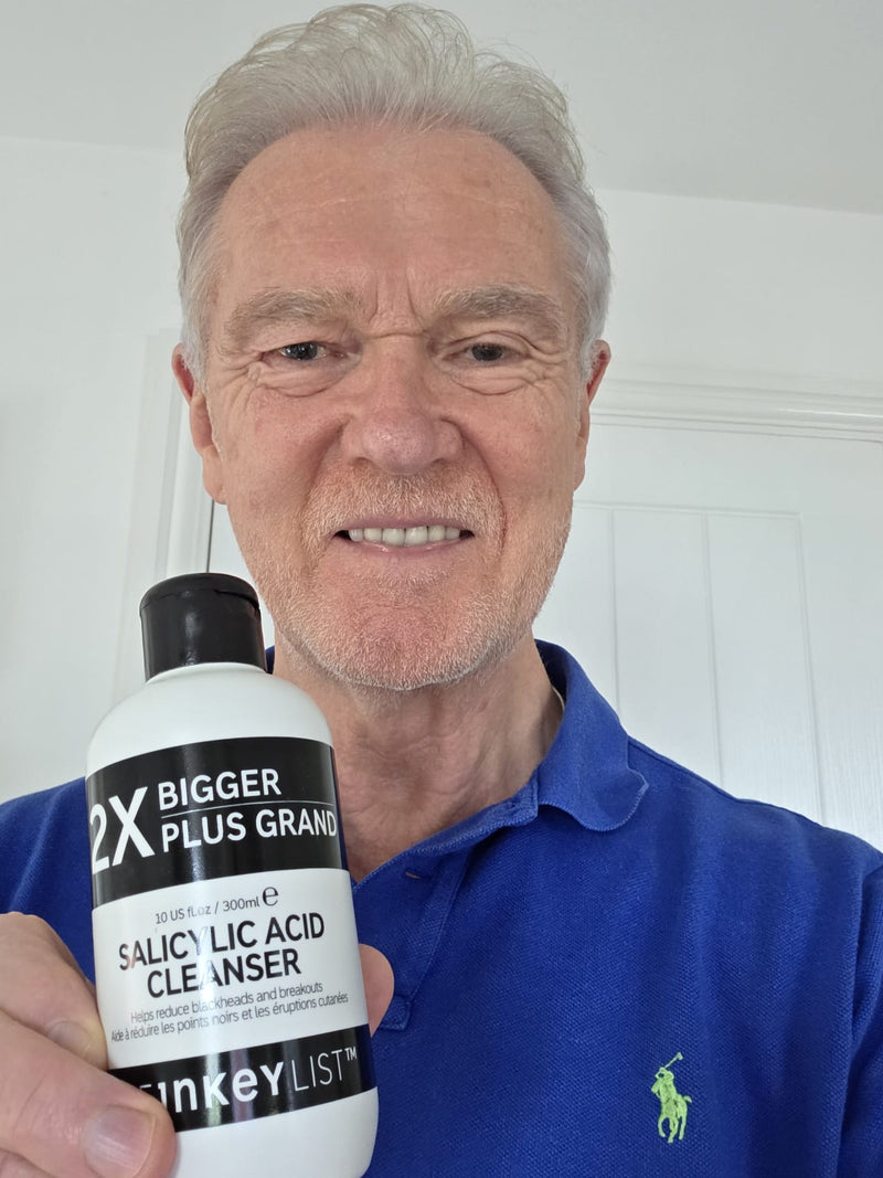 An older man with gray hair and a beard wearing a blue polo shirt with a green embroidered polo player logo, holding up a white bottle of '2X Salicylic Acid Cleanser' by 'The Inkey List'.