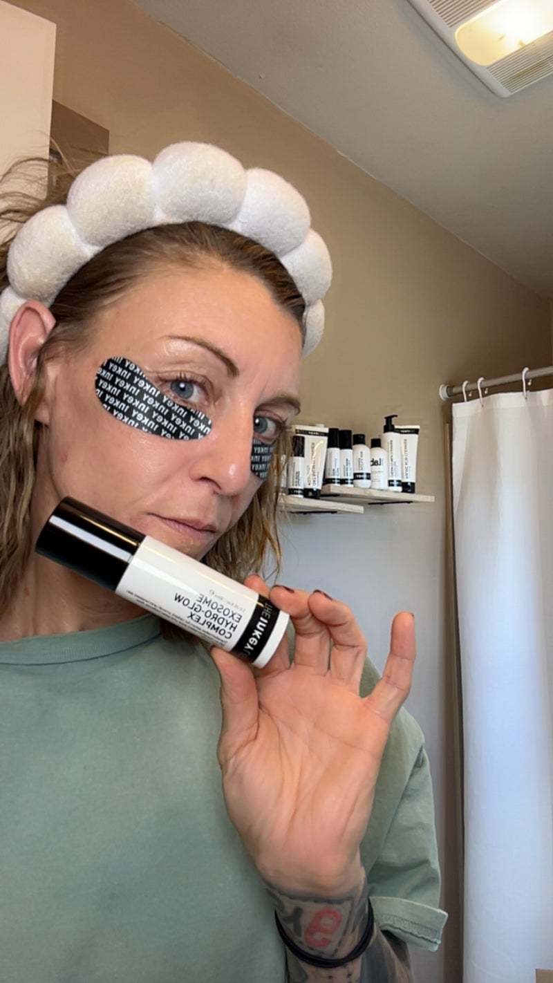 A woman in a bathroom wearing a white pom-pom headband and eye tape, holding a black and white cosmetic tube near her face.