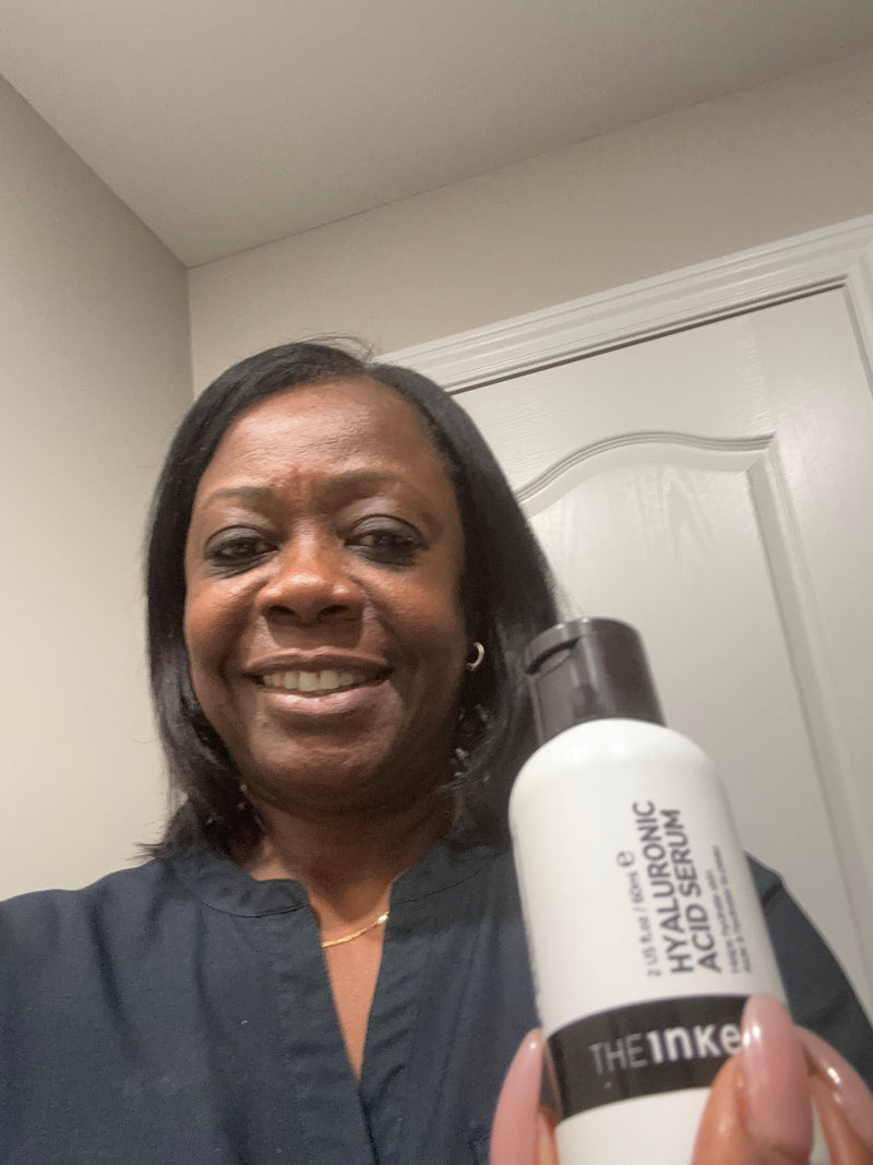 A woman holding a white bottle of The Inked brand Hyaluronic Acid Serum in front of a white door and beige wall.