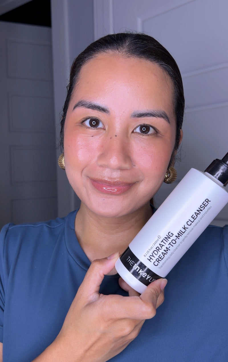 A woman holding a white bottle of Hydrating Cream-to-Milk Cleanser against a dark gray background.