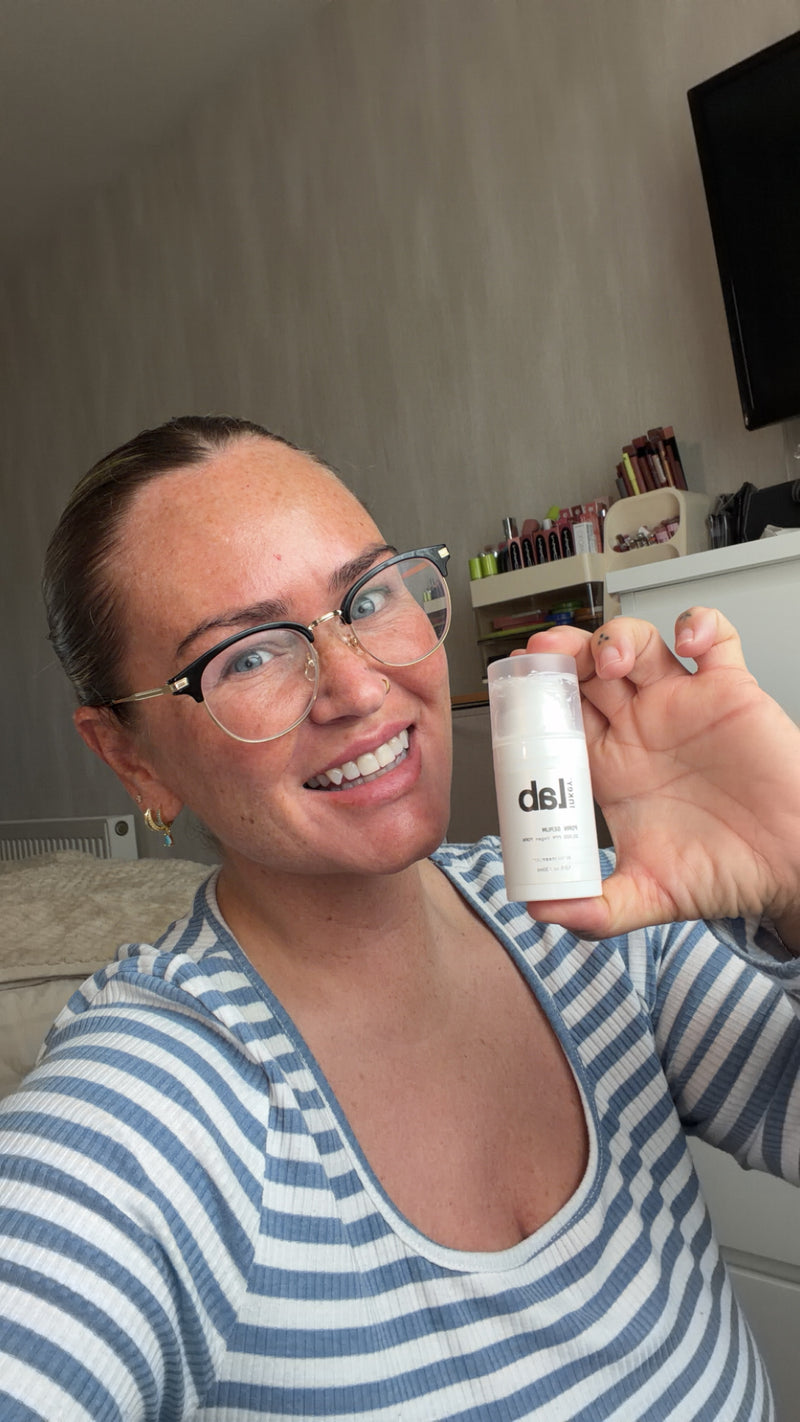A woman wearing glasses and a blue and white striped shirt holds up a small white bottle with the brand name 'PDRN' visible on the front, in a home setting.