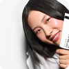 A close-up lifestyle shot of a woman with long black hair holding a white tube of oat balm next to her face against a clean white background.
